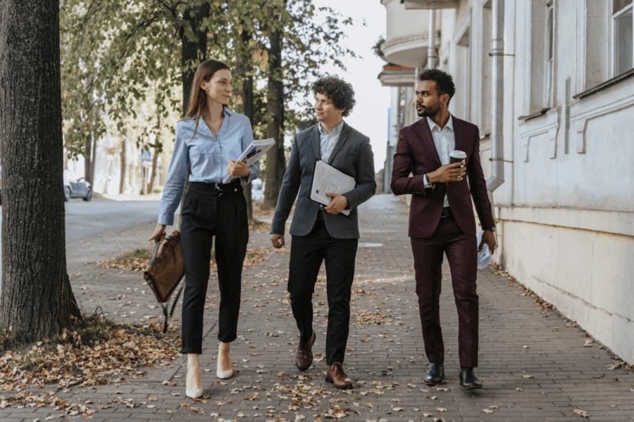 Three professionals walking on a tree-lined sidewalk, discussing with folders and coffee in hand, conveying a collaborative and focused mood.